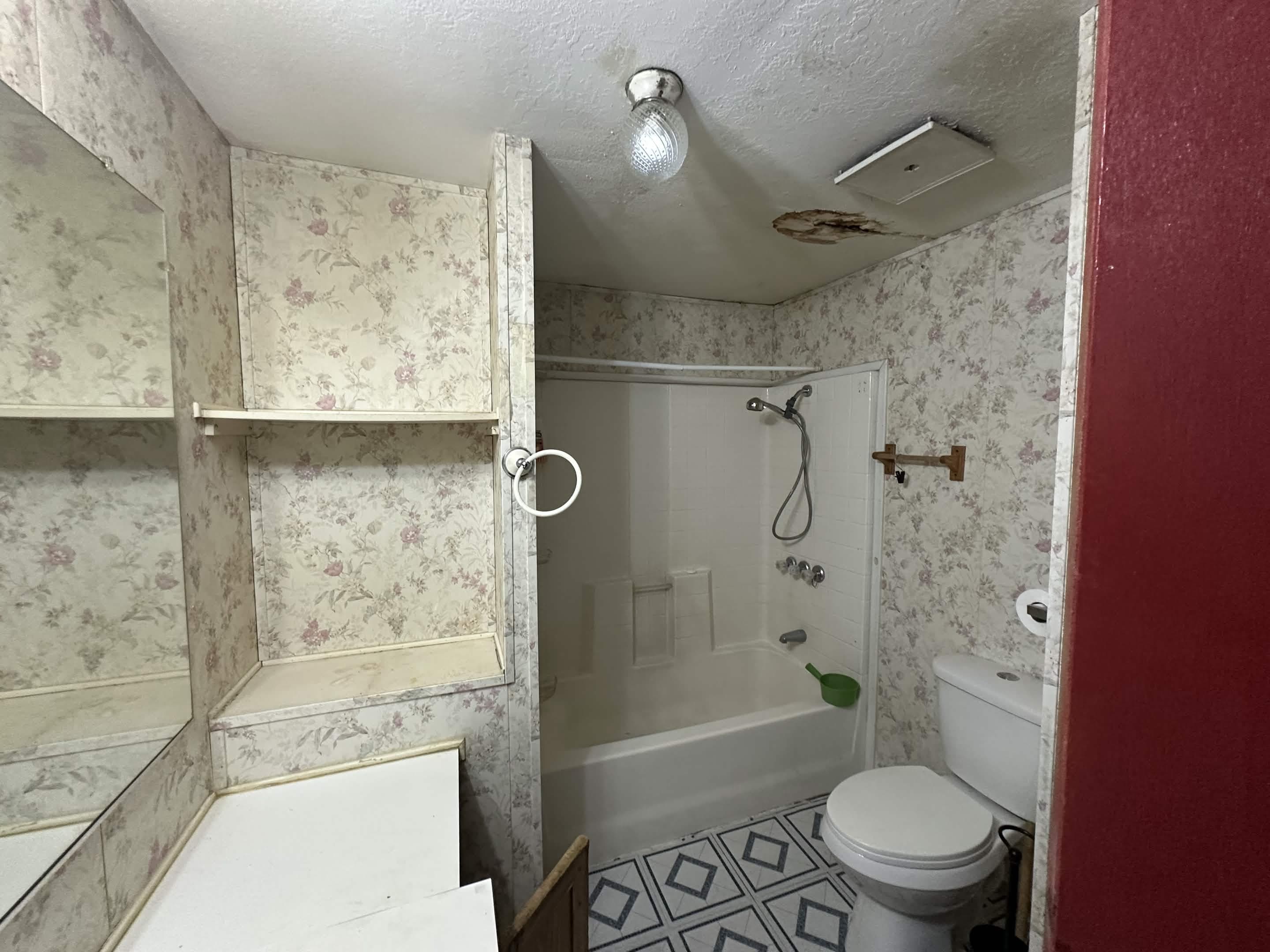 Bathroom before remodel — dated floral wallpaper, old tub, and water-damaged ceiling